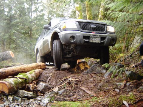 1985 Ford Bronco - A dark-colored SUV is navigating a challenging off-road course, with its front right tire elevated over rocks and logs in a muddy forest se…