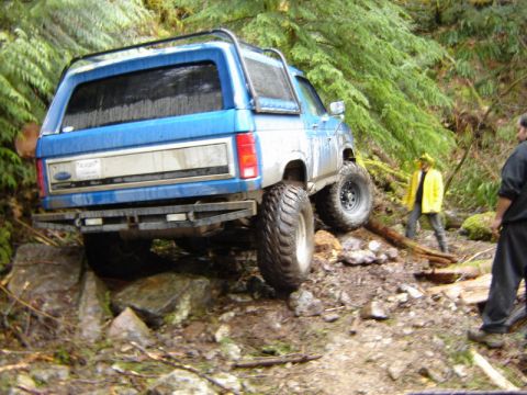 1985 Ford Bronco - A blue Ford Bronco is navigating a rocky, wooded trail. The rear of the SUV is angled towards the viewer as it drives over large rocks and…