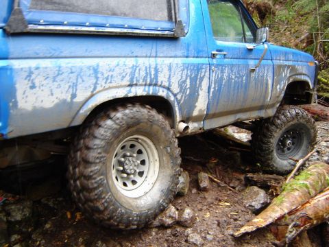1985 Ford Bronco - A blue Ford Bronco is shown from the side, navigating a muddy, wooded trail with large off-road tires. The vehicle is heavily splattered wi…