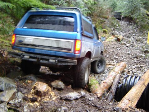1985 Ford Bronco - A blue Ford Bronco is navigating a rough, wooded trail, driving over rocks and logs. Another vehicle is visible further up the trail.