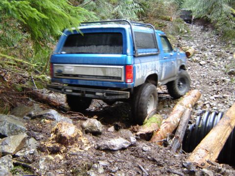 1985 Ford Bronco - A blue Ford Bronco is navigating a muddy, rocky off-road trail with fallen logs and a culvert. Another vehicle is visible further up the tr…