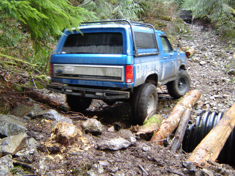 Blue Ford Bronco driving on a muddy, rocky off-road trail with logs and a culvert.