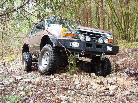1985 Ford Bronco - A lifted, modified brown SUV is navigating a rocky, wooded trail, showcasing its aggressive off-road tires and custom front bumper.
