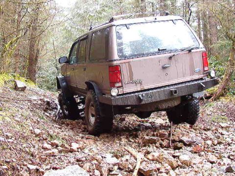 1985 Ford Bronco - A brown SUV is navigating a rocky, wooded trail with its tires in a muddy rut. The rear of the vehicle is angled away from the camera, show…