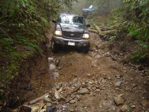 1985 Ford Bronco - A dark-colored Ford pickup truck navigates a muddy, rocky off-road trail through a dense forest. Another blue off-road vehicle is visible f…