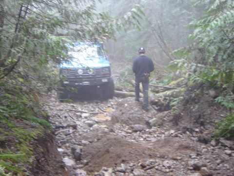 1985 Ford Bronco - A blue Ford Bronco drives down a rocky, muddy trail in a foggy forest. A person wearing a dark long-sleeved shirt and a hat walks behind th…