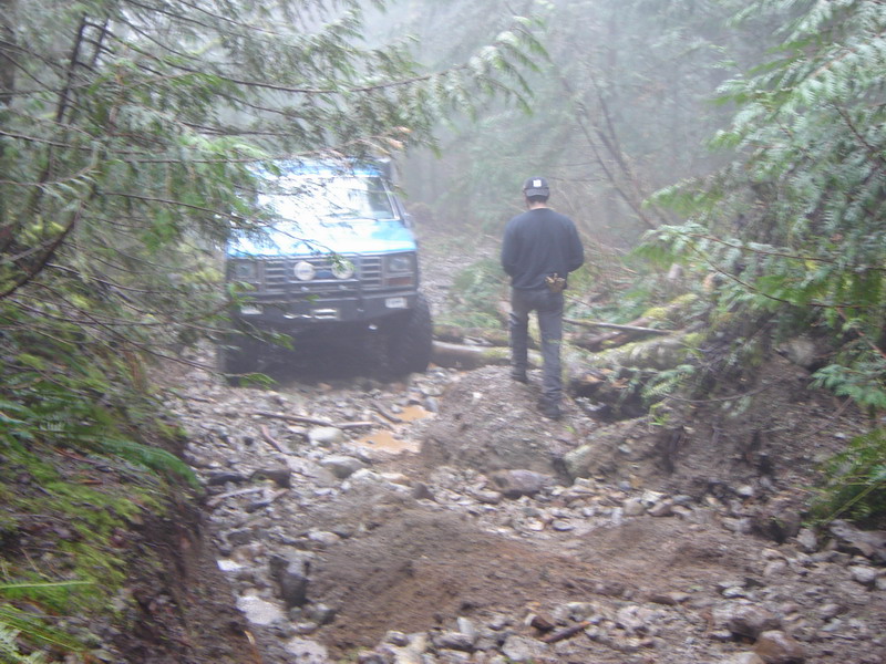 Blue Ford Bronco on a rocky forest trail with a person walking behind it.