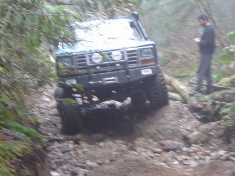 1985 Ford Bronco - A blue 1980s Ford Bronco is navigating a rocky, off-road trail in a wooded area. A person stands in the background, looking at their phone.