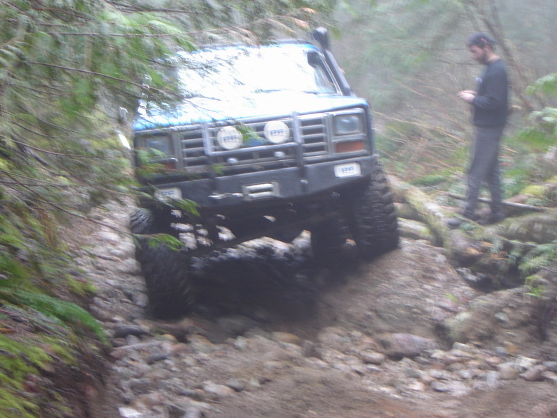 Blue 1980s Ford Bronco off-roading on a rocky trail in a forest.
