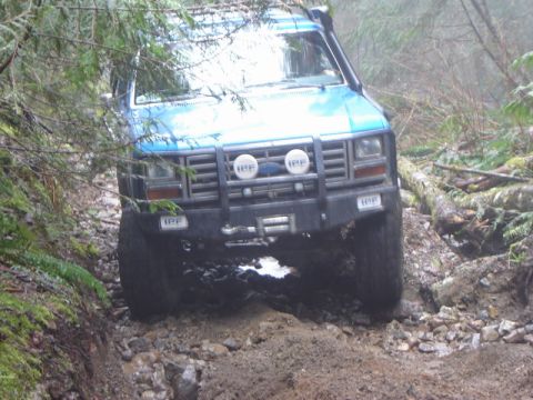 1985 Ford Bronco - A blue Ford Bronco is navigating a rocky, wooded trail. The vehicle has off-road modifications including IPF lights and a snorkel.