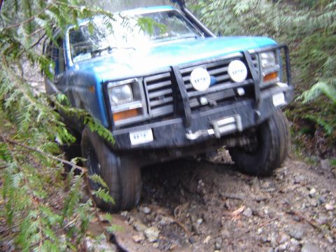 1985 Ford Bronco - A blue Ford Bronco is shown navigating a muddy, wooded trail, with its front end angled towards the camera and branches obscuring the view.