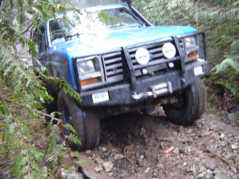 Blue Ford Bronco driving on a muddy, wooded trail.