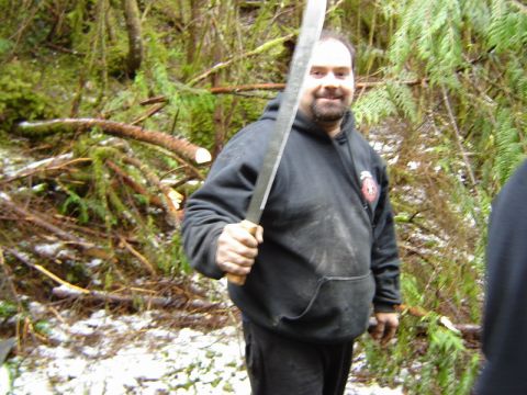 1985 Ford Bronco - A man in a black hoodie holds a large machete in a wooded area with snow on the ground.