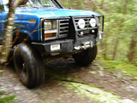 1985 Ford Bronco - The front quarter of a blue Ford Bronco is shown off-road, with large tires and a custom bumper with auxiliary lights and a winch. The vehi…
