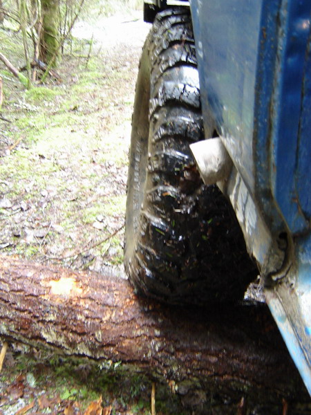 1985 Ford Bronco - A close-up, low-angle view of the right rear tire of a blue Ford Bronco driving over a fallen log in a wooded area. The tire is caked in mu…