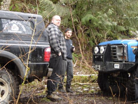 1985 Ford Bronco - Two men stand next to a black pickup truck and a blue off-road vehicle in a wooded area with wet ground. The black truck has a "North Shore…