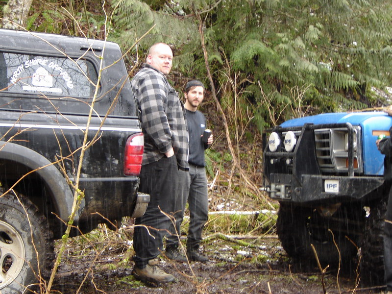 Two men stand by a black pickup truck and a blue off-road vehicle in a wooded area.