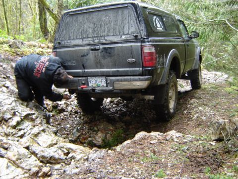 1985 Ford Bronco - Rear 3/4 view of a mud-caked dark green Ford 4x4 pickup with a color-matched canopy, climbing a rocky, root-strewn trail in a wet forest. A…