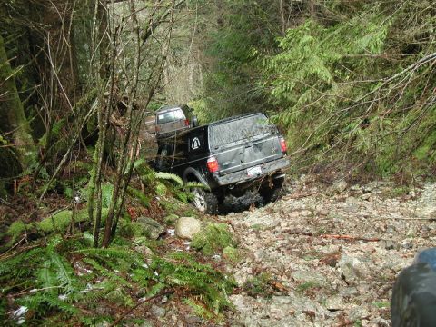 1985 Ford Bronco - Two off-road pickup trucks navigate a rocky, wooded trail. The lead truck is black with a cap, and the second truck is brown.