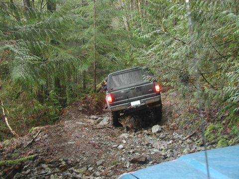 1985 Ford Bronco - A black pickup truck with a camper shell navigates a rocky, muddy trail through a dense forest.