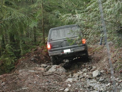1985 Ford Bronco - A dark-colored pickup truck with a camper shell is navigating a rocky, wooded trail. The truck is angled uphill, with its rear taillights i…