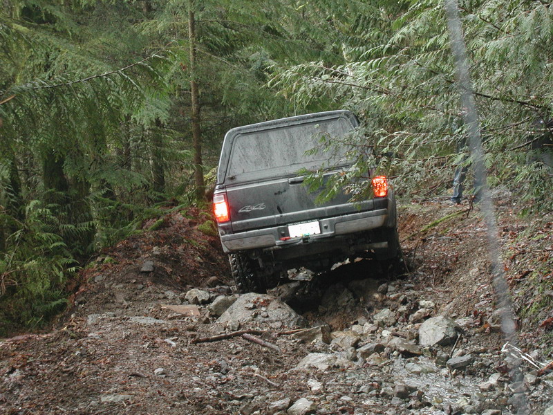 Dark pickup truck with camper shell driving on a rocky, wooded trail.