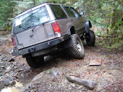 1985 Ford Bronco - A lifted, muddy brown Ford Explorer is navigating a steep, wooded trail with large off-road tires. The rear of the SUV is angled upwards, s…