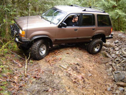 1985 Ford Bronco - A brown, lifted Ford Explorer is navigating a muddy, rocky trail in a forest. The driver is visible looking out the window.
