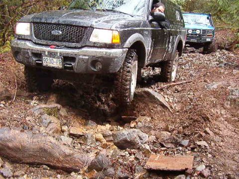 1985 Ford Bronco - A black Ford Ranger is navigating a rocky, muddy off-road trail, with a blue Ford Bronco following behind. The Ranger's front license plate…