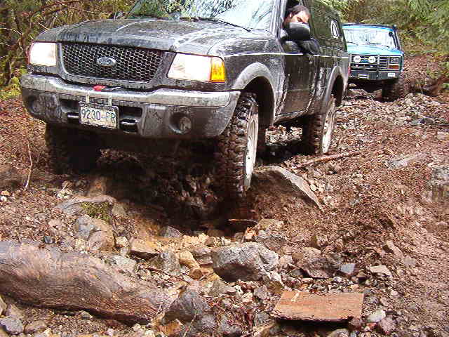 Black Ford Ranger driving on a rocky, muddy off-road trail, blue Ford Bronco behind.
