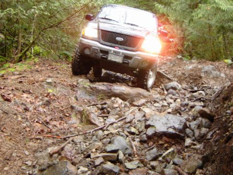 1985 Ford Bronco - A dark-colored Ford pickup truck with its headlights on navigates a rocky, wooded trail. The truck is angled towards the viewer, showing it…