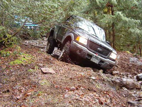 1985 Ford Bronco - A dark-colored Ford pickup truck is navigating a steep, muddy, and rocky off-road trail. The truck is tilted to the side as its tires grip…
