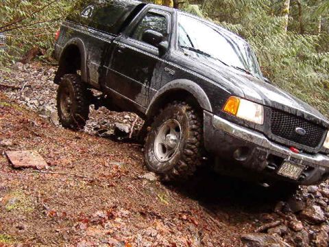 1985 Ford Bronco - A black Ford Ranger pickup truck with off-road tires is navigating a steep, muddy, and rocky incline in a wooded area.
