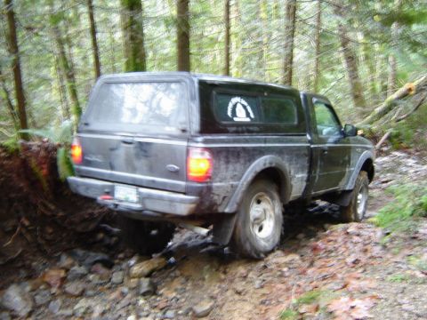 1985 Ford Bronco - A dark-colored pickup truck with a camper shell is navigating a muddy, rocky trail through a forest. The truck is angled to the right, with…