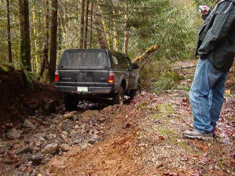 1985 Ford Bronco - A black Ford pickup truck with a camper shell is navigating a muddy, rocky trail in a forest. A person in jeans and a jacket stands to the…