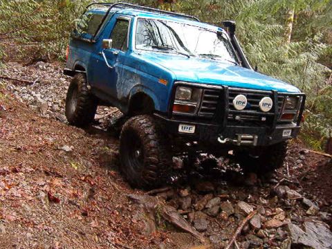 1985 Ford Bronco - A blue 1980s Ford Bronco is navigating a steep, rocky, and muddy off-road trail with a snorkel visible on the driver's side. The vehicle is…