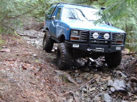 1985 Ford Bronco - A blue Ford Bronco is navigating a rocky, wooded trail, with its large tires gripping the uneven terrain. The front of the vehicle features…