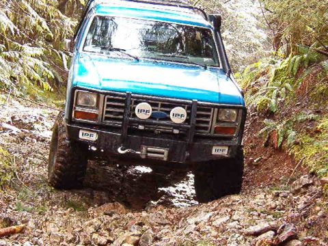 1985 Ford Bronco - A blue 1985 Ford Bronco is shown from a low front angle, navigating a muddy, rocky trail in a wooded area. The vehicle features a brush gua…