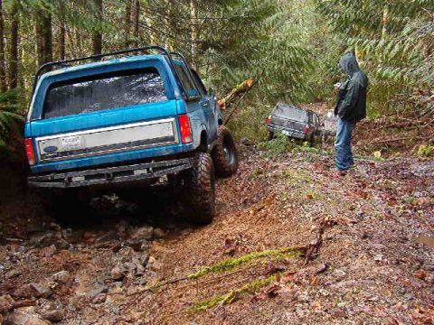 1985 Ford Bronco - A blue Ford Bronco is parked on a muddy, wooded trail with another SUV in the background. A person in a hooded jacket stands to the right o…