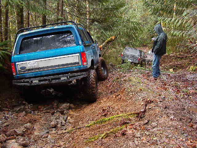 Blue Ford Bronco on a muddy forest trail with another SUV and a person.