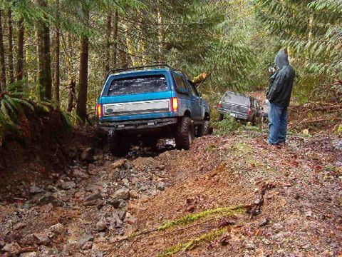 1985 Ford Bronco - A blue Ford Bronco is navigating a muddy, rocky trail through a dense forest, with another dark SUV visible further up the path. A person i…