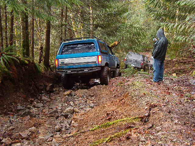 Blue Ford Bronco on a muddy forest trail with another SUV behind it.
