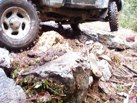 1985 Ford Bronco - The front tire of an off-road vehicle is shown navigating a rocky and muddy terrain. The vehicle appears to be a truck or SUV, with its und…