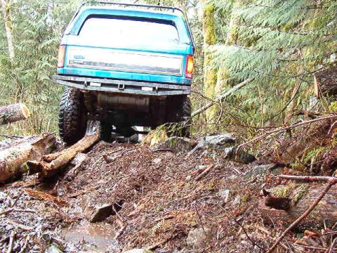 1985 Ford Bronco - The rear of a blue Ford Bronco is shown navigating a rough, wooded trail with large logs and debris on the ground.