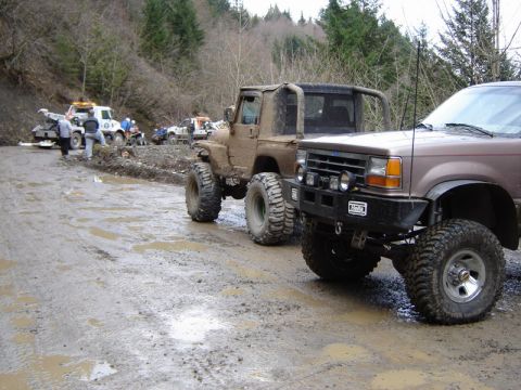 1985 Ford Bronco - Several off-road vehicles, including a lifted Ford Explorer and a muddy Jeep, are parked on a wet, muddy road in a wooded area. A tow truck…