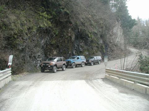 1985 Ford Bronco - Three off-road vehicles are parked on a gravel road next to a steep, rocky embankment. The vehicles are a brown SUV, a blue SUV, and a blac…
