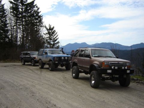 1985 Ford Bronco - A line of four modified off-road vehicles, including a brown SUV and a blue Bronco, are parked on a dirt road with a mountainous forest in…
