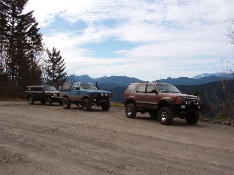 1985 Ford Bronco - Three modified off-road vehicles are parked on a dirt road with a mountainous landscape in the background. The vehicles include a black pic…