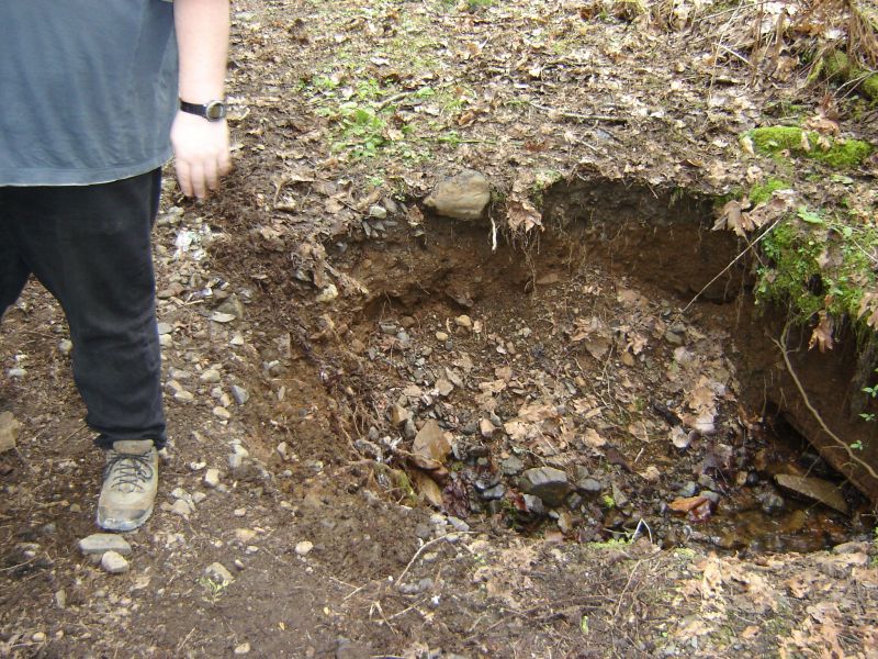 Person standing next to a hole in a dirt embankment with a small stream at the bottom.