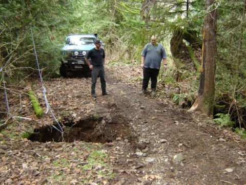 1985 Ford Bronco - Two men stand on a dirt path next to a large hole in the ground, with a blue off-road vehicle parked behind them in a wooded area.